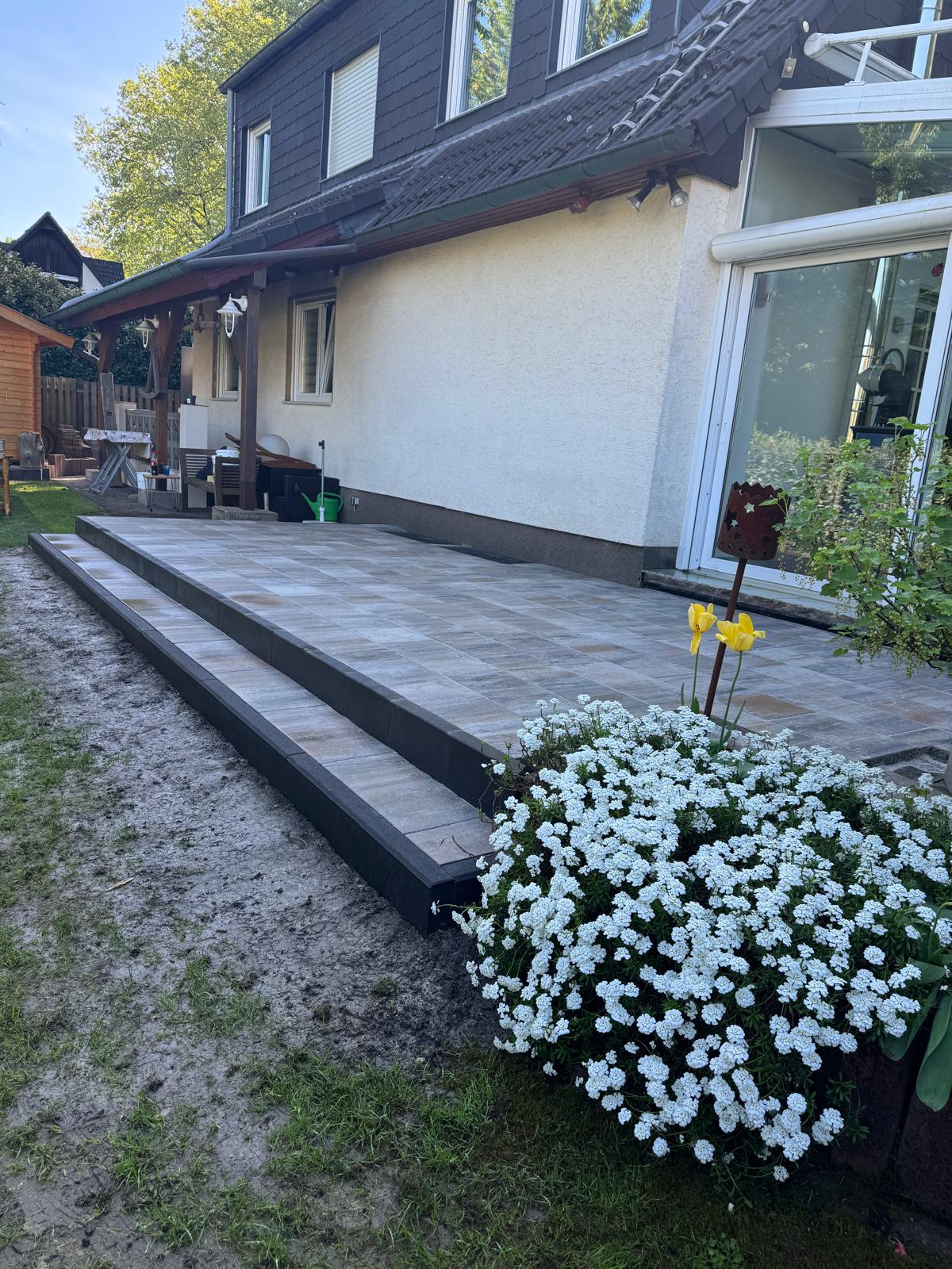 Eine Terrasse mit Fliesen, weißen Blumen im Vordergrund und gelbem Blütenstecker